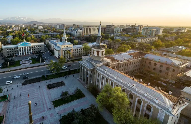 Aerial view of Bishkek city skyline with historical buildings and distant mountains at sunset.