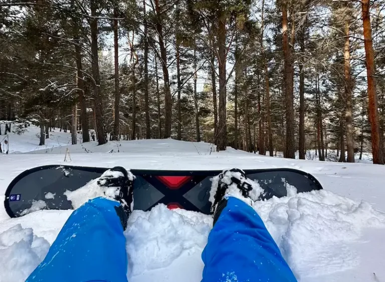 Snowboarder's point of view looking down at a board with snow-covered pine trees at Karakol ski resort